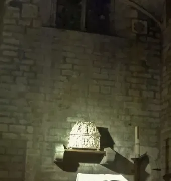 Ornately carved Sant Cugat casket with relief scenes, resting on a wooden shelf against a stone wall.