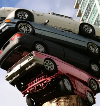 Trans Am Rapture sculpture with stacked cars rising from a large tree trunk against a blue sky, with a modern building in the background.
