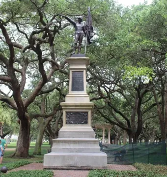 The Defenders of Fort Moultrie sculpture atop a stone plinth in a park, with a figure in military attire raising one arm and holding a flag.