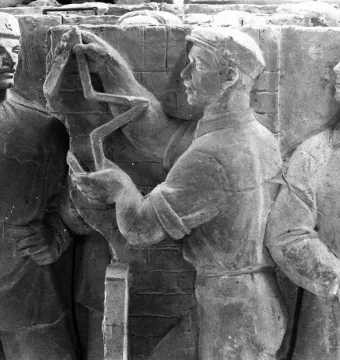 Weathered stone relief with three male figures in caps and uniforms around a central lightning-bolt symbol.