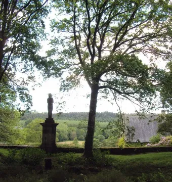 The Vénus de Quinipily stone sculpture stands on a tall pedestal between two trees, with fields and forest in the background.