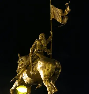 Golden Joan of Arc equestrian statue seen from behind-left, with a flowing banner and bright light beneath the horse.