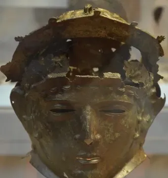 Ribchester Helmet with a patinated bronze face mask and damaged upper section on a display stand.