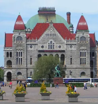 Aleksis Kivi Memorial, a green-patinated seated statue on a stone pedestal in a city square with a large stone building behind it.