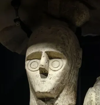 Weathered stone head from the Giants of Mont\'e Prama with concentric circle eyes and a simple mouth, mounted on metal supports.