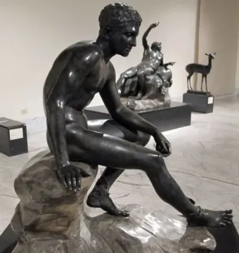 Seated Hermes bronze statue of a nude male figure on a rock, looking downward.