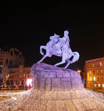 The Monument to Bohdan Khmelnytsky lit in purple, with a bright cluster of white lights at its base in a nighttime city square.