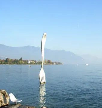 Fork of Vevey sculpture standing in the water of Lake Geneva near a rocky shore, with mountains and buildings in the distance.