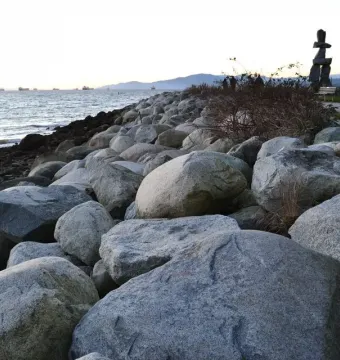 An Inukshuk stands near a rocky shoreline beside calm water at twilight, with mountains and ships in the distance.