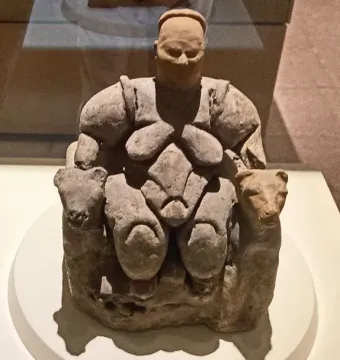 Seated Woman of Çatalhöyük figurine with animal heads on the armrests, displayed in a museum case.