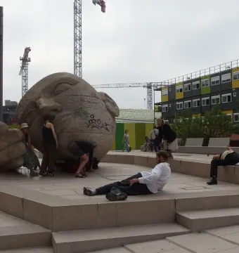 Écoute sculpture showing a large stone head lying on its side beside a cupped hand, with people around it in a plaza.