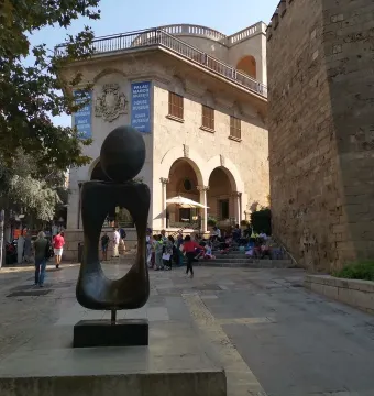 Monument a la dona, a dark abstract bronze sculpture on a plinth in a paved plaza with historic buildings behind it.