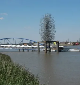 Quantum Cloud sculpture on a riverside platform beside a blue pedestrian bridge.
