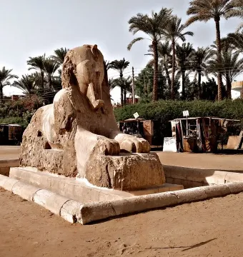 The Sphinx of Memphis on a low stone base in a sandy open area with palm trees and small stalls in the background.
