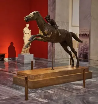 The Jockey of Artemision bronze sculpture shown with a horse and young rider on a pedestal in a museum gallery.