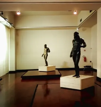 Two Riace bronzes standing on pedestals in a museum gallery.