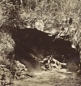 Sepia photograph of the cave entrance at Brassempouy, with a man seated in the dark opening surrounded by vegetation.