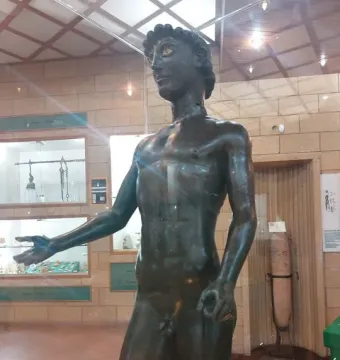 Efebo de Selinunte, a dark bronze youthful nude male statue with one arm extended in a museum display.