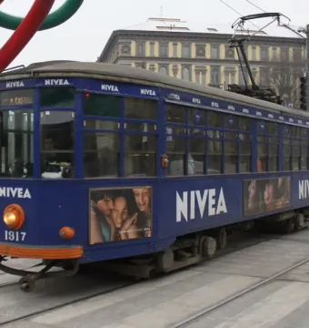 A blue NIVEA tram passes street tracks with parts of the Needle, Thread and Knot sculpture visible above.