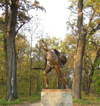 Bronze Greek Warrior Monument of a helmeted figure holding a shield and short blade beside tall trees.