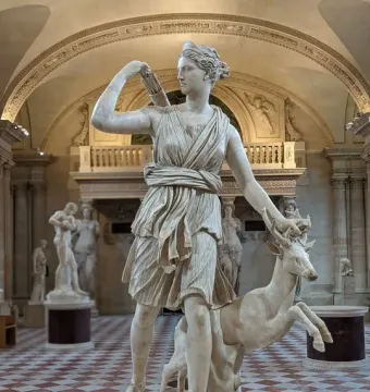 Diana of Versailles, a marble statue of a woman reaching back toward a quiver while resting her hand on a deer’s antlers in a museum hall.