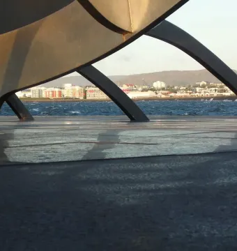 The Sun Voyager sculpture seen through its curved metallic forms, with blue water, a distant cityscape, and hills in the background.
