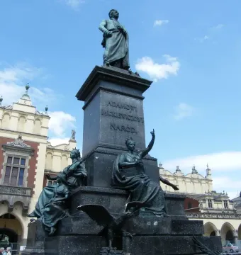 Adam Mickiewicz Monument with a bronze standing figure on a tall pedestal, framed by historic buildings and a blue sky.