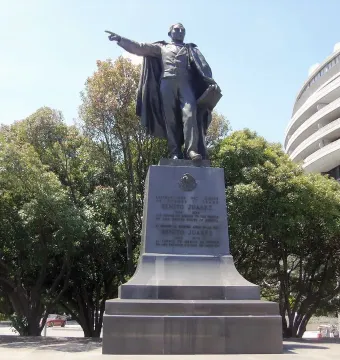 Bronze statue of Benito Juarez standing on a tall inscribed pedestal with trees and a curved building in the background.