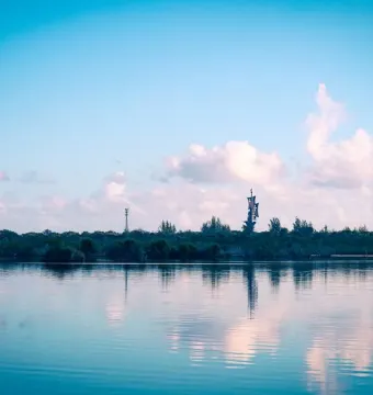Birth of the New World sculpture reflected in calm water near a tree-lined shore.