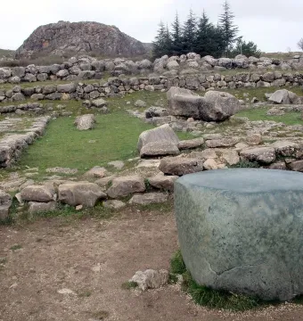 Large greenish stone block in the foreground at Hattusa, with ruins of stone walls and a rocky hill behind it.