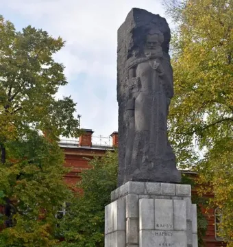 Karl Marx Monument on a tiered stone base with Cyrillic inscriptions, set among trees and a red brick building.