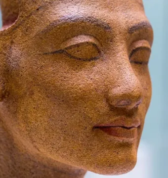 Right-side close-up of the unfinished head of Nefertiti with carved facial features and an ear against a blurred blue background.