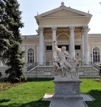 White marble Laocoön sculpture standing on a pedestal in front of a museum building, framed by trees and a lawn.