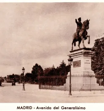 Marqués del Duero monument beside a wide tree-lined avenue in Madrid, with a fence and buildings in the background.