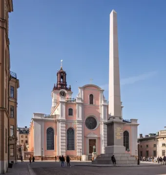 The Obelisk at Slottsbacken stands on a dark tiered pedestal beside a pink and white church with a clock tower.