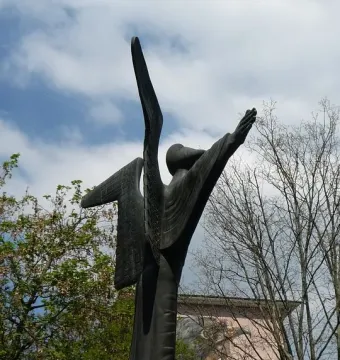 Angel of Peace sculpture with raised arms against a cloudy sky, standing among trees near a mural-covered building.