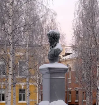 Frans Michael Franzén statue on a snow-covered pedestal in Oulu, with leafless trees and buildings behind it.
