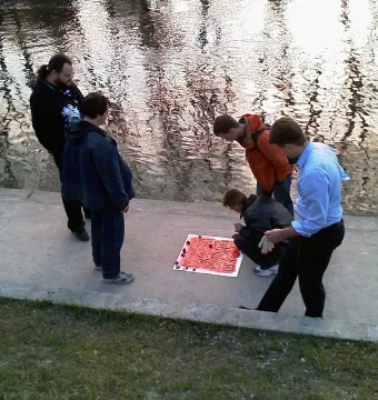 Five men stand beside the Keyboard Monument, looking at a large red panel with raised square blocks laid on a concrete path near the water.