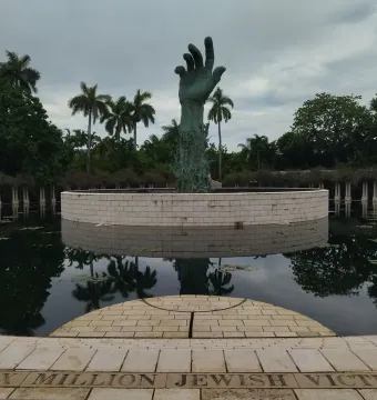 The Holocaust Memorial sculpture in Miami Beach shows a verdigris arm and hand rising from a circular stone base in a reflecting pool, with palm trees in the background.