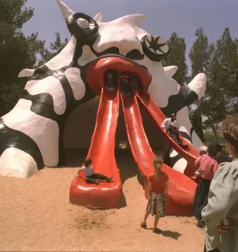The Golem monster slide sculpture with three red chutes, with children playing in a sandy park.