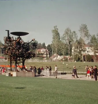 Heureka kinetic sculpture standing on a lawn with visitors nearby and trees in the background.