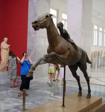 The Jockey of Artemision bronze sculpture displayed in a museum, showing a rearing horse with a small rider leaning forward.