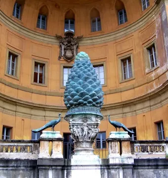 La Pigna pine cone sculpture on an ornate pedestal, flanked by two peacock statues in front of a curved orange building.