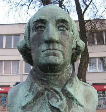 Weathered green-patinated bust of George Washington on a speckled stone base, with a building and bare branches behind it.