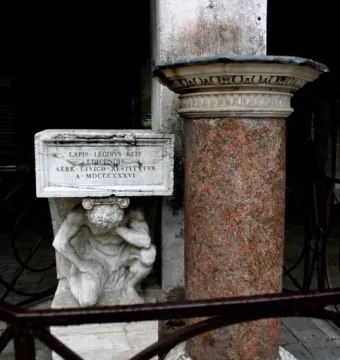 Il Gobbo di Rialto, a white marble crouching figure holding up an inscribed plaque beside a reddish-brown column.