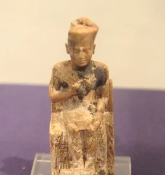 Khufu statuette of a seated ivory figure with a tall crown, shown on a clear base.