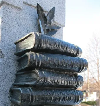 La mort de Dante monument detail showing four stacked dark metal books mounted on a stone pillar, with leafy decoration above the top volume.