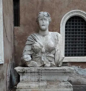 Madama Lucrezia, a weathered stone female statue with draped garments and an eroded face against a reddish-brown wall.