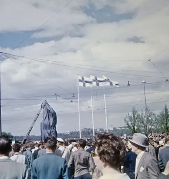 Crowd gathered around the covered Marshal Mannerheim equestrian statue during its unveiling, with flags overhead and a ladder beside it.