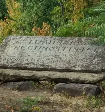 Weathered stone Thanksgiving Candle monument with a carved inscription on a tilted rectangular slab, set among rough stones and greenery.